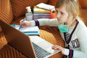 Young white female student with a mask doing school work at home on laptop and couch
